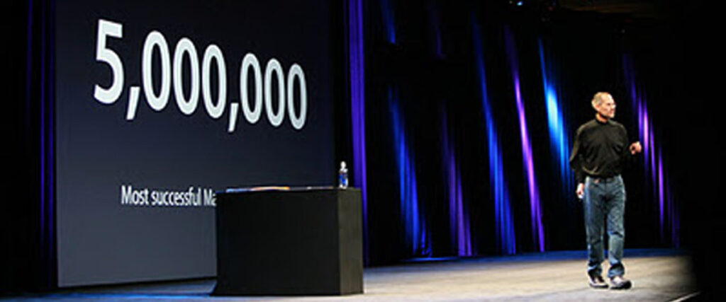 A person in a black turtleneck, blue jeans, and sneakers stands on a stage beside a podium with a water bottle on it. Behind them, the screen displays "5,000,000" with the text "Most successful" beneath it during their presentation.