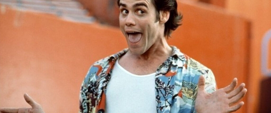 A person with dark hair is wearing a colorful, tropical-themed shirt over a white tank top. They are looking at the camera with a wide, open-mouthed smile and raised eyebrows, with both hands spread wide and fingers splayed. The background is orange, making it perfect for a lively presentation slide template.