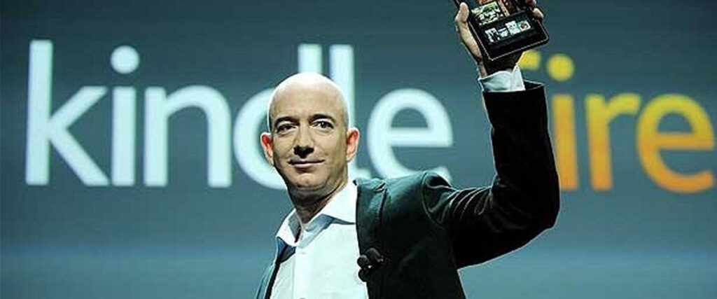 A person in a suit holds up a Kindle Fire device proudly on stage during a presentation. The background has the words "kindle" and partially visible "fire" in large letters.