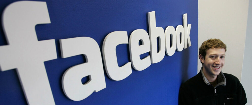 A person stands in front of a large "facebook" logo on a blue wall, smiling, as if ready to add the final touch to their presentation.
