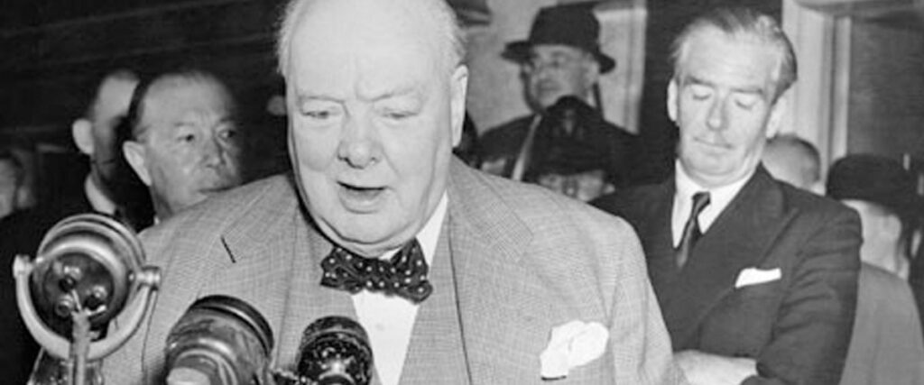 A black-and-white photograph captures an elderly man in a suit speaking into a microphone, surrounded by people. The man has a balding head and is wearing a bow tie. A man in the background, standing with arms folded, looks down as if contemplating the presentation.