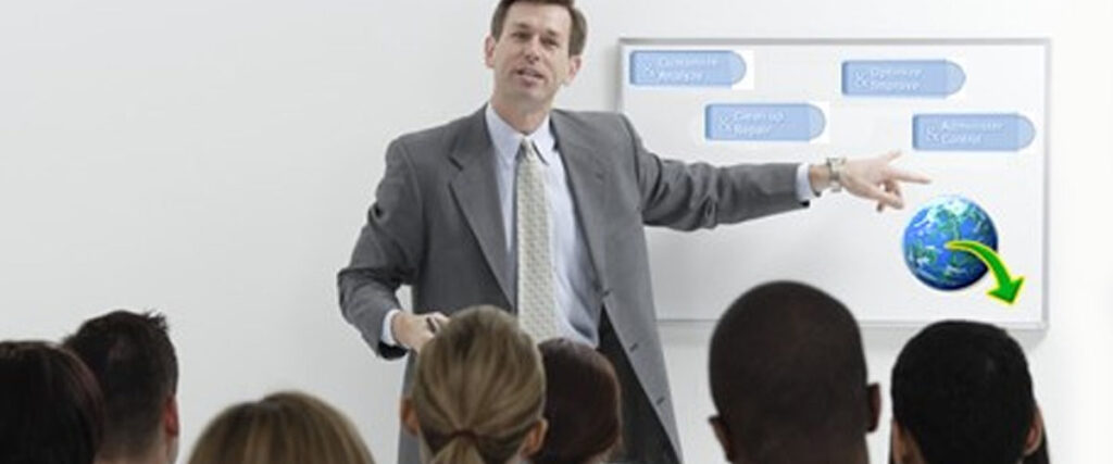 A man in a suit is giving a presentation in front of an audience. He points to a whiteboard with organizational charts and a slide that shows the Earth with a downward arrow. The audience is seated and attentively facing him.