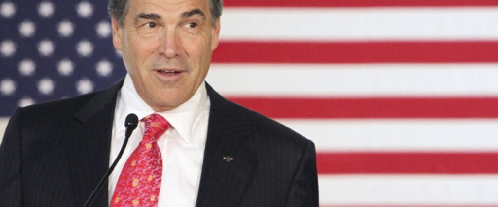 A man in a dark suit and red tie delivers a serious presentation into a microphone, standing in front of an American flag backdrop.