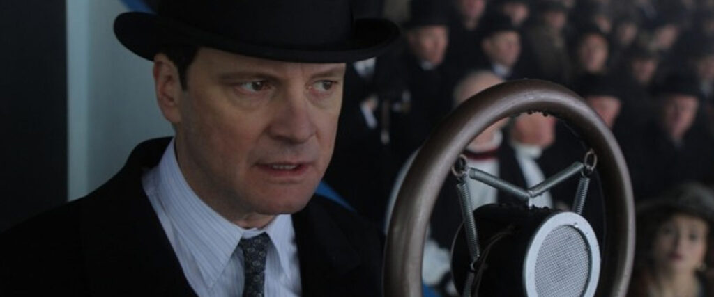 A man in a suit and bowler hat speaks into an old-fashioned microphone. He is surrounded by blurry onlookers in dark clothing. His face is tense and focused, suggesting he is delivering an important pitch deck presentation.