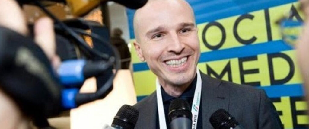 A smiling bald man in a suit and tie stands before an array of microphones, speaking to the press. In the background, there's a large sign with partially visible text that reads "SOCIAL MEDIA." The scene appears to be a media interview or press conference, perfect for inclusion in a PowerPoint slide.