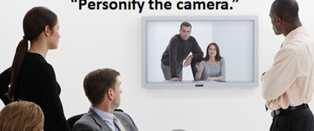 A group of five people in an office setting participate in a video conference. Three individuals are physically present and watching a large screen on which two other colleagues are visible. The text above the screen reads, "Personify the camera," as they work on their pitch deck together.
