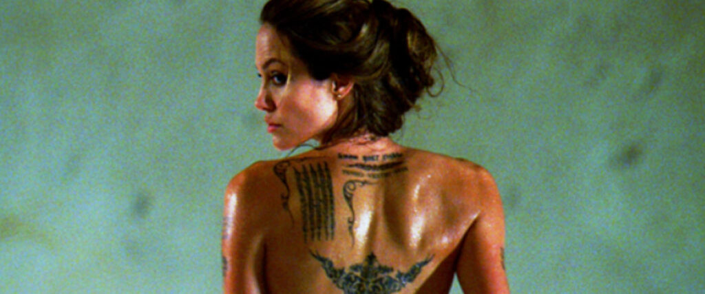 A person with dark hair tied up looks over their shoulder, revealing several intricate tattoos on their upper back. The background is blurred and neutral, giving a sleek appearance perfect for a pitch deck or slide presentation. The person's skin appears slightly glistening.