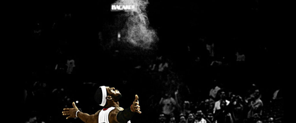A basketball player stands on a court with arms outstretched and head tilted back, surrounded by a cloud of white powder. The background is dimly lit, with spectators faintly visible in the stands. A sign overhead reads "INCANTE," reminiscent of a dynamic slide template for an electrifying presentation.