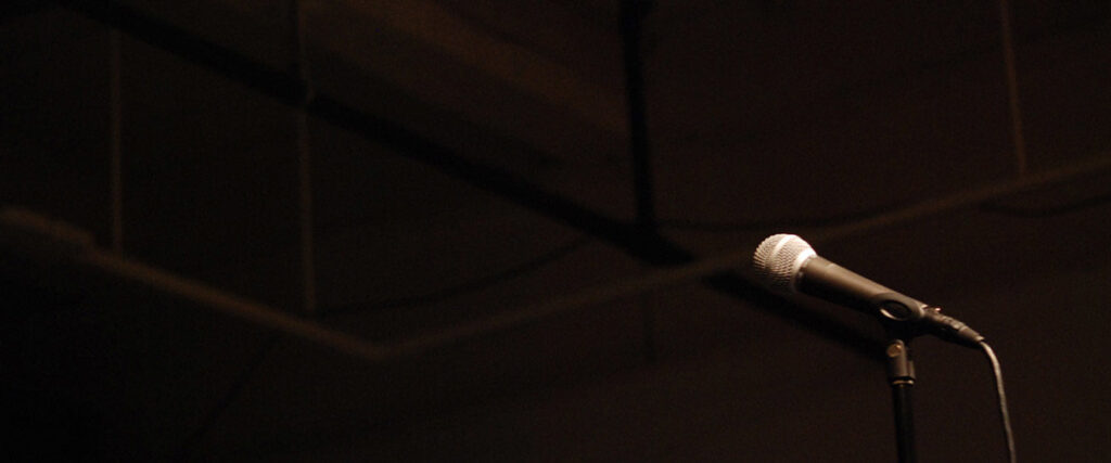 A microphone is mounted on a stand against a dark background, possibly on a stage or in a studio. The scene is dimly lit, casting subtle shadows and emphasizing the microphone as the focal point. Perfect for use in a PowerPoint slide template to create an impactful presentation moment.