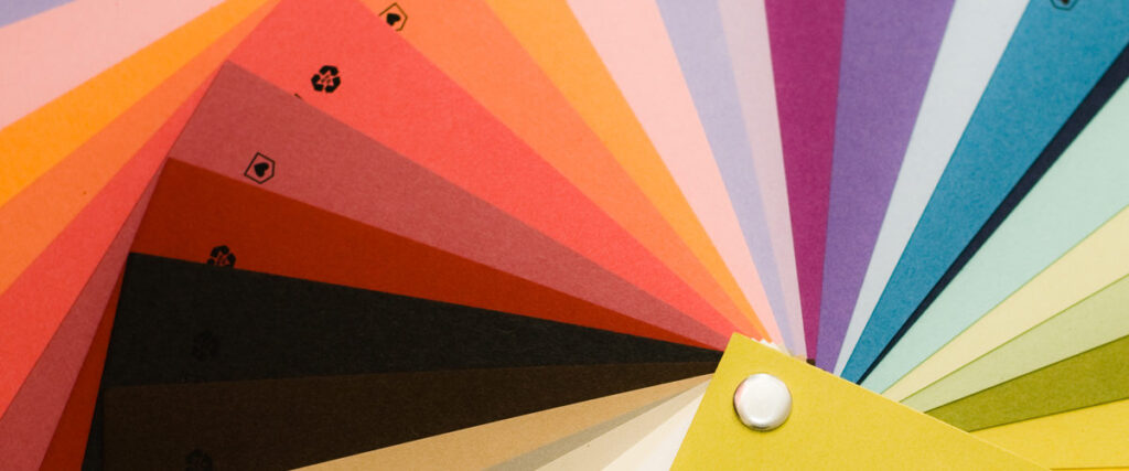 A fanned-out collection of colorful paper swatches arranged in a gradient. The swatches range from warm colors like reds and yellows to cool colors like blues and purples. Each swatch is held together by a metal rivet in the corner, making it perfect for use in a PowerPoint slide or pitch deck.