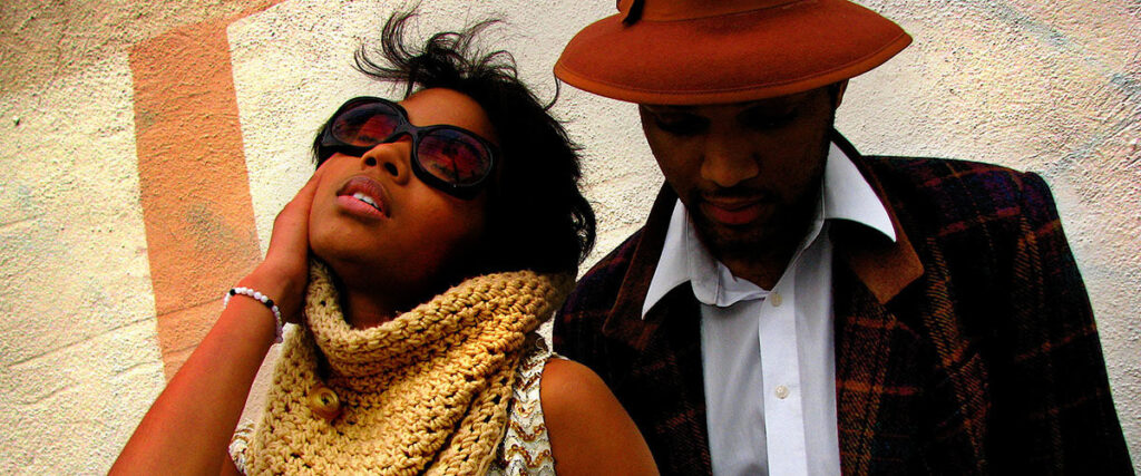 A woman wearing large sunglasses and a knitted scarf poses with her hand on her face, while a man in a plaid jacket and brown hat looks downward. They stand against a textured wall with subtle graphic designs, creating an aesthetic that could inspire your next PowerPoint presentation.