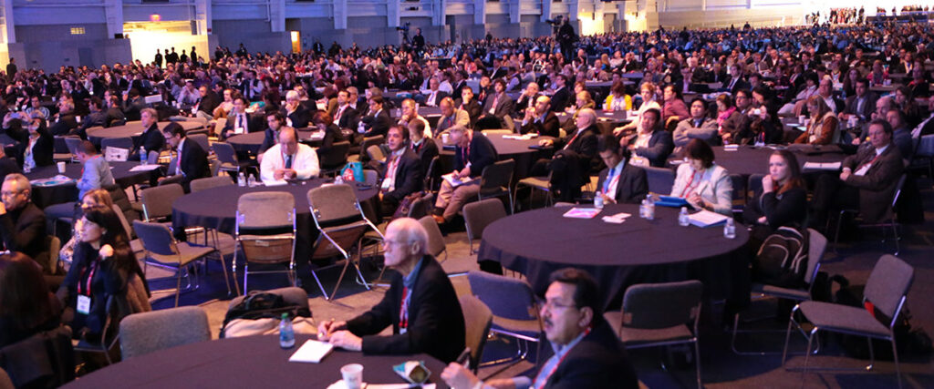A large gathering of people seated at round tables in a spacious conference hall, attentively listening to a speaker. The audience consists of men and women in professional attire, with some individuals taking notes or looking at their electronic devices. A PowerPoint presentation is displayed on a screen behind the speaker.