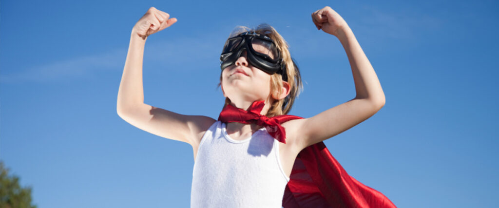 A child wearing aviator goggles and a red cape stands against a clear blue sky, flexing their biceps in a superhero pose. The child has a white tank top on and appears to be imagining themselves as a superhero, ready to conquer the world. This image perfectly captures the essence of inspiration for your next PowerPoint pitch deck.