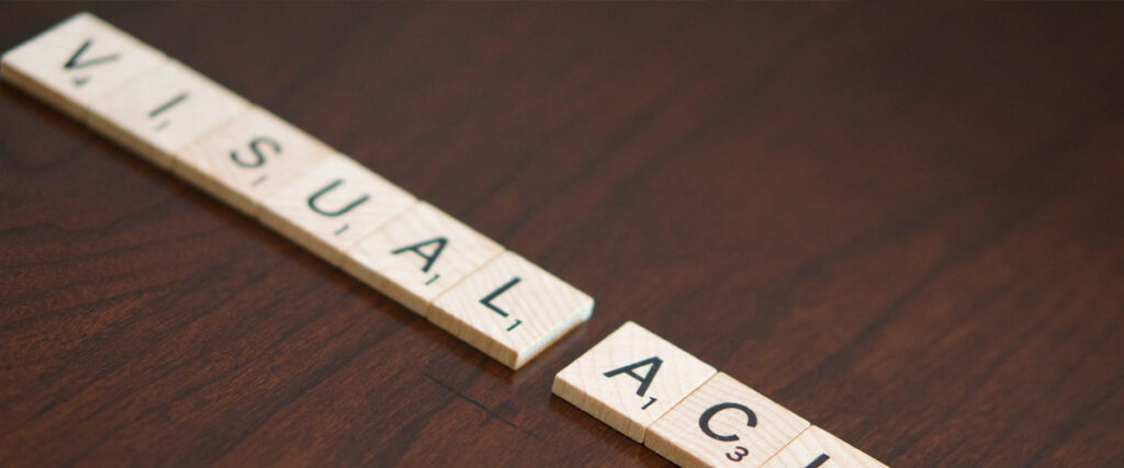 Scrabble tiles spelling out "visual" and "acm" are arranged against a wooden surface background, evoking the meticulous craft of designing a compelling presentation or pitch deck.