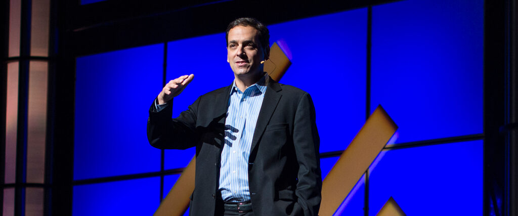 A man in a dark suit and light blue shirt gestures while delivering a pitch deck on stage, with a backdrop of a blue screen featuring yellow geometric shapes. The stage is lit with bright lights.