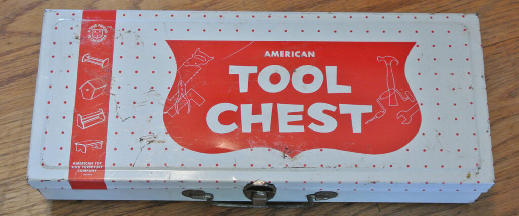 A rectangular, white metal box with red graphics and the words "American Tool Chest" written on it. The box has a polka dot pattern and illustrations of tools around the text. Slightly scuffed, it has a latch on the front. This could be a great addition to your presentation slide on tool storage solutions.