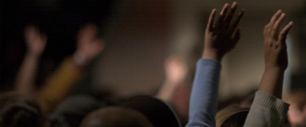 Several raised hands reaching upwards dominate the foreground, with a blurred background suggesting a crowd or audience in a dimly lit setting. This dynamic scene can be effectively used in a PowerPoint slide to emphasize participation and engagement.