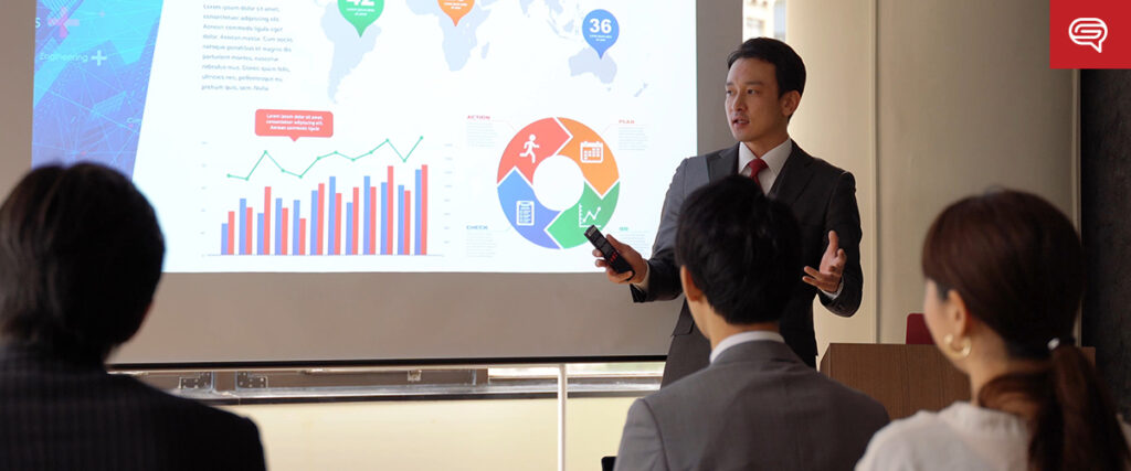 A business professional in a suit is presenting a pitch deck to a small audience. The presentation screen displays colorful charts, graphs, and world maps. The audience includes both men and women, seated and attentively listening.