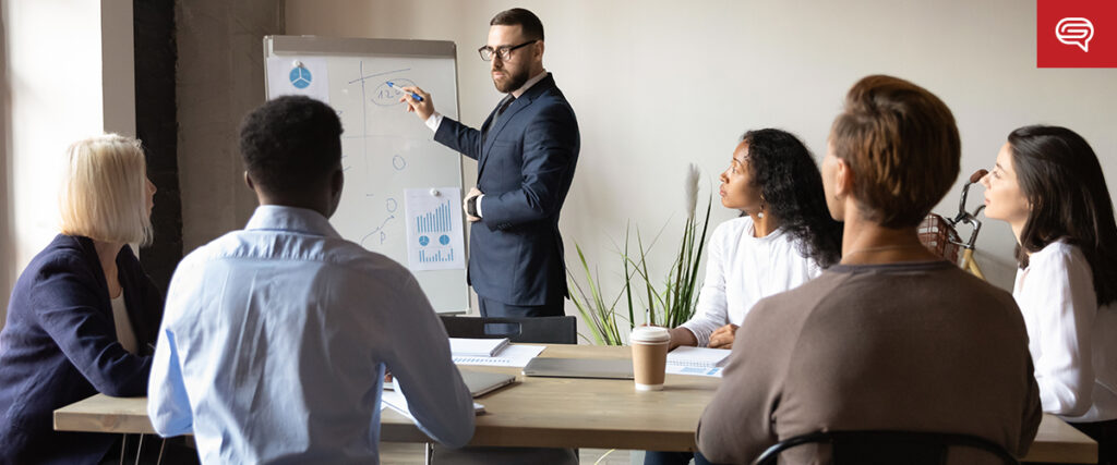 A man in a suit stands at the front of a meeting room, pointing at a flip chart filled with diagrams. Five people, three women and two men, sit around a table listening attentively to the presentation. Documents, notebooks, and a coffee cup are on the table.