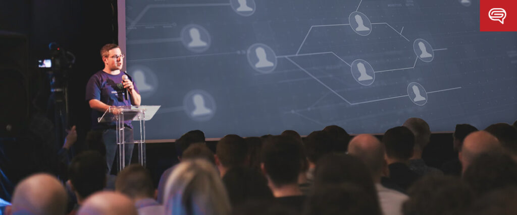 A person wearing glasses and a dark shirt speaks into a microphone at a clear podium. Behind them is a large screen displaying a PowerPoint slide template of connected user icons. An audience is seated in front, attentively listening. A red logo is in the top-right corner.