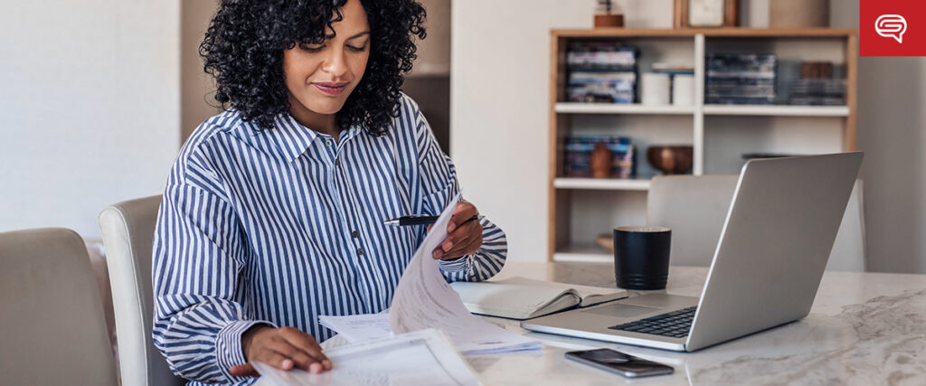A woman with curly hair is sitting at a table, holding a pen and looking at documents. She is wearing a striped shirt and working on a laptop, possibly designing a PowerPoint slide template. A smartphone and notebook are on the table next to a coffee mug, with a bookshelf in the background.