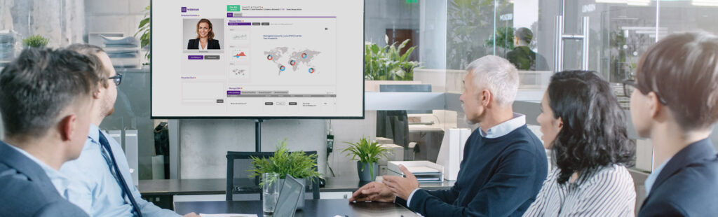 Five people in business attire sit at a conference table, facing a large screen displaying charts, graphs, and a world map. A pitch deck with a woman’s profile picture and details is also visible on the screen. The setting is a modern office with glass walls and potted plants.