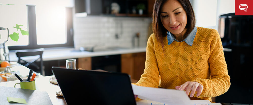 A woman with shoulder-length hair is sitting at a kitchen table, wearing a yellow sweater. She is looking at papers in her hand while working on a laptop, possibly reviewing a pitch deck. The kitchen in the background has a modern design with light-colored walls and a window letting in natural light.