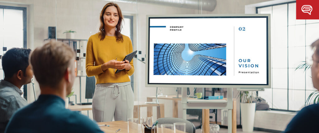 A woman in a yellow sweater presents a company profile pitch deck to three seated colleagues in a modern office. The slide on the screen reads "Our Vision" and features an abstract architectural design. The room exudes a professional atmosphere.