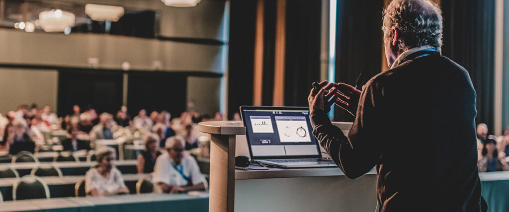 A man is speaking at a conference, standing behind a podium with a laptop displaying PowerPoint presentation slides. Attendees sit in rows, facing the stage in a large, well-lit room. The speaker's back is to the camera.