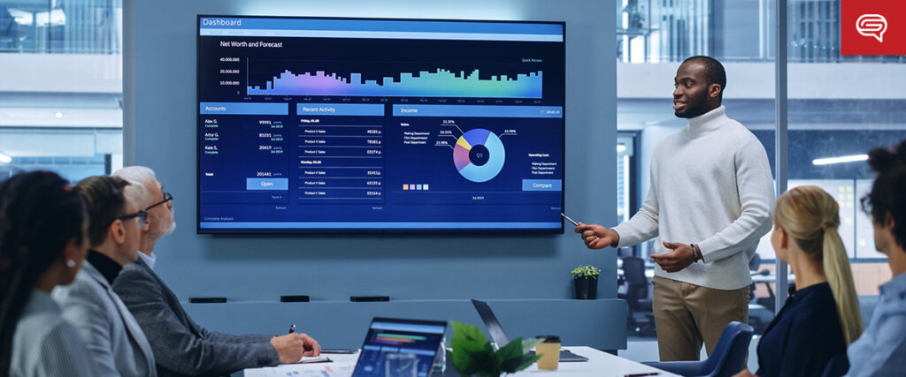 A man is standing and presenting data on a large screen to a group of people in a modern office meeting room. The PowerPoint slides display various charts and graphs, including a bar chart, line graph, and pie chart. Attendees are seated around a table with laptops.