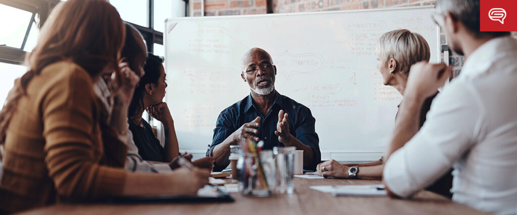 A group of six people having a meeting in a modern office setting. A bald man in the center, wearing glasses and a dark shirt, is speaking passionately while pointing at a slide on the screen. The group members listen attentively, and a whiteboard with diagrams and notes is visible in the background.