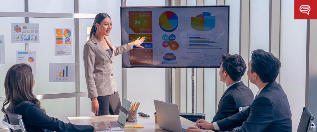 A woman in a business suit is presenting a PowerPoint slideshow with charts and graphs on a large screen to three seated colleagues in an office setting. There are additional charts on the glass walls behind her. Everyone is engaged and focused on the presentation.
