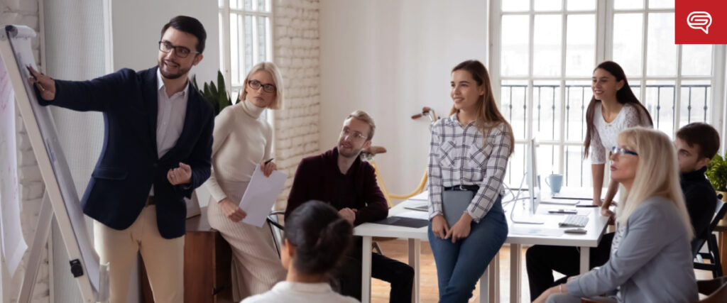 A diverse group of seven professionals in a modern office setting are engaged in a presentation. One man, in a blazer, stands next to a flip chart pointing at it while others sit and stand around a table, attentively listening and taking notes. The slide he is discussing captures everyone’s focus.