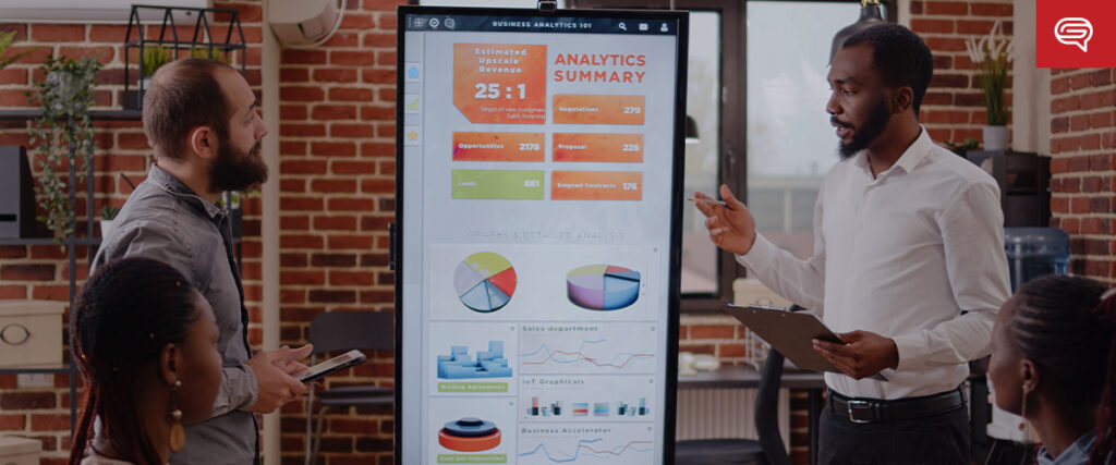 A man presents a PowerPoint slide on a large screen while another man listens, holding a tablet. They stand in a modern office with brick walls and a glass door. Three other individuals are seated, listening and looking at the screen, some taking notes.