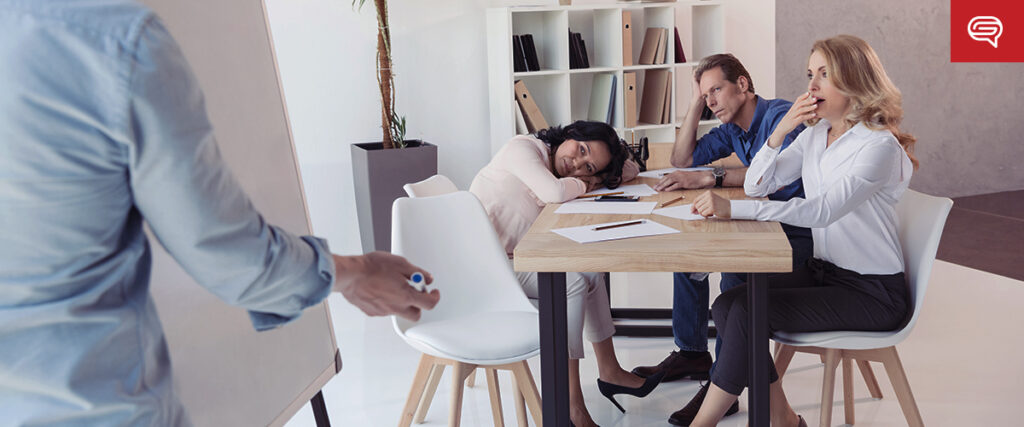 Three colleagues sit wearily at a table during a meeting, two of them appearing bored or tired, with one resting their head on their arms. A fourth person stands in front of a flipchart, conducting the presentation using a pitch deck. The mood is lethargic and inattentive.