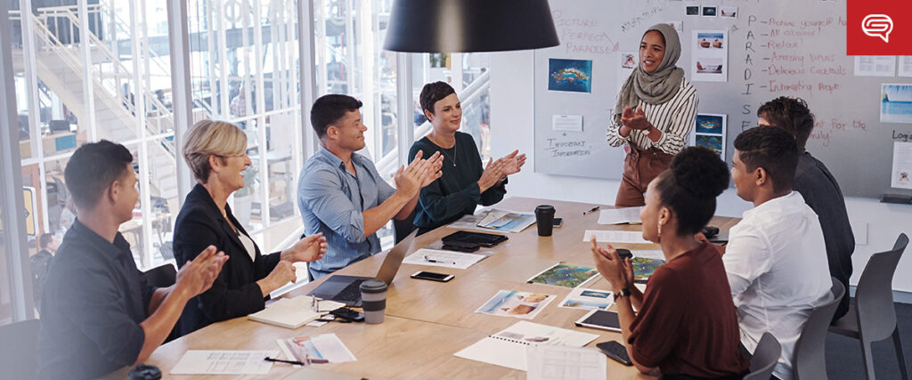 A diverse group of professionals sits around a conference table, clapping as a woman in a hijab stands and smiles. The room has large windows, a whiteboard with notes, and various documents and devices on the table. It seems to be a business presentation featuring an engaging slide template.