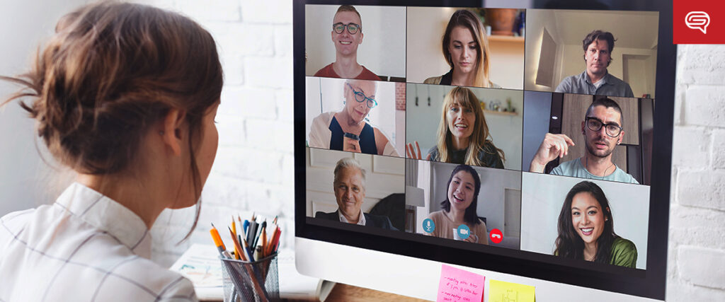 A woman sits at a desk with colorful sticky notes and pens, attending a video conference on her monitor. The screen shows nine people in various indoor settings, engaging in the virtual meeting, likely discussing a PowerPoint presentation.