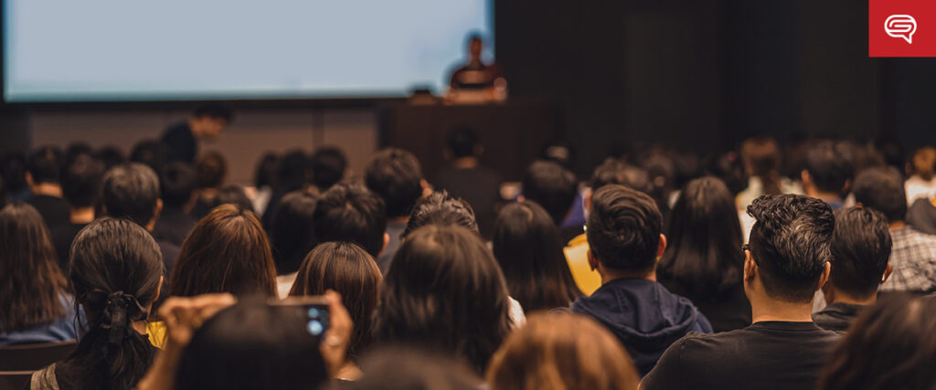 A room full of people sit and face a stage where a person is giving a presentation. The audience is attentive, their backs to the camera. The room is dimly lit, and the PowerPoint slide template on the screen is visible but not readable.