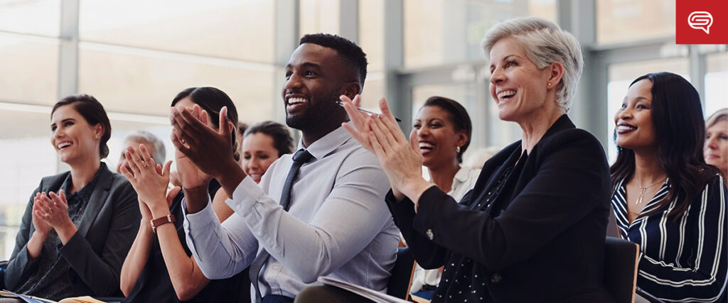 A diverse group of professionals, including men and women of various ages, are sitting in a well-lit conference room, smiling and applauding during a PowerPoint presentation. Some are dressed in business attire. Large windows in the background let in natural light.