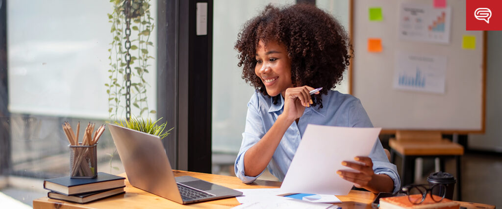 A woman with curly hair sits at a desk by a window, smiling as she looks at a paper in her hand. A laptop, a cup, and office supplies are on the desk. A whiteboard with charts and graphs, resembling slide templates for her pitch deck, is in the background.