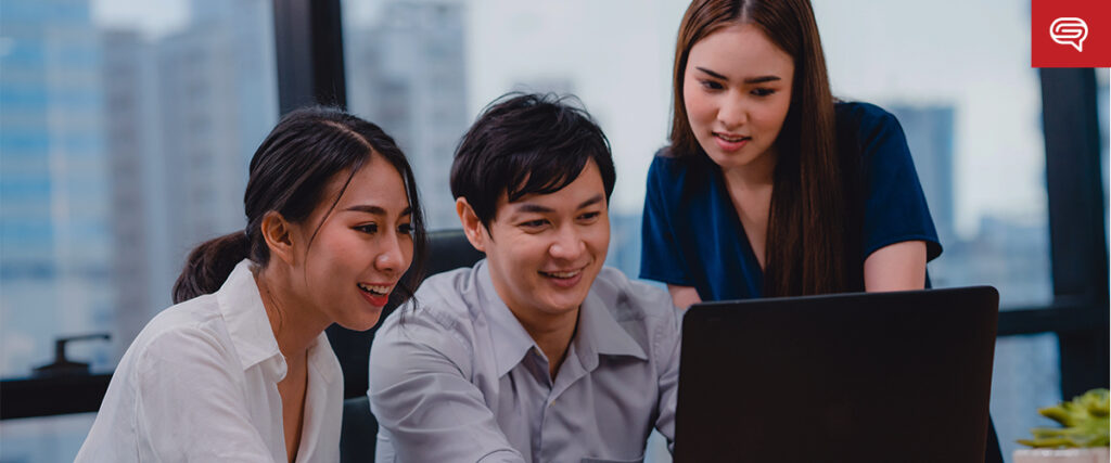 Three people in an office workspace are gathered around a laptop, engaged in a discussion. The background shows large windows with a view of city buildings. They appear focused and collaborative, possibly reviewing a presentation slide template.