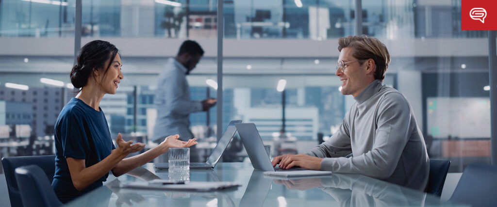 Two people are seated at a glass conference table with laptops, engaged in a conversation. A third person in the background is standing and looking at a tablet. The office environment is modern with large windows and a busy backdrop; they seem to be discussing their latest pitch deck.