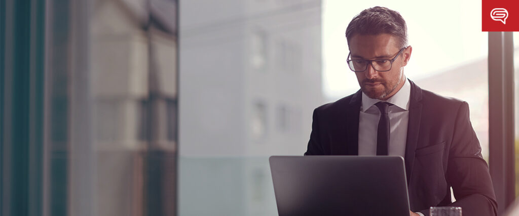 A man wearing a suit and glasses is focused on his laptop while seated in an office setting with large windows. The natural light from the windows illuminates the space, and a partially visible red speech bubble logo is seen in the upper right corner as he reviews his pitch deck.