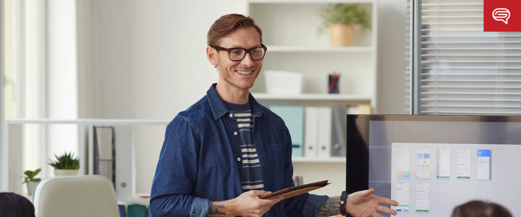 A man wearing glasses and a blue shirt holds a tablet while presenting to a small group. Behind him, a screen displays a slide template of user interface design. Shelves with decorative plants and office supplies are visible in the background.