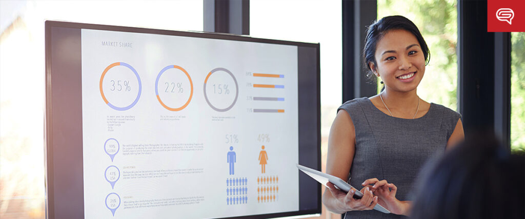 A woman stands next to a large screen displaying various charts and graphs about market share percentages. Holding a tablet, she smiles as she presents information to an unseen audience. The setting is a modern office with large windows, emphasizing her professional pitch deck presentation.