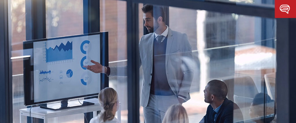A man in a business suit stands and gestures toward a screen displaying charts and graphs on a presentation slide. Three people are seated around a table, attentively watching. The room has large windows with a view outside.