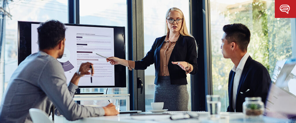 A woman in business attire stands at a large screen displaying a slide presentation while speaking to two seated colleagues in a modern office. She gestures towards the screen, and sunlight streams through the windows, illuminating the scene.