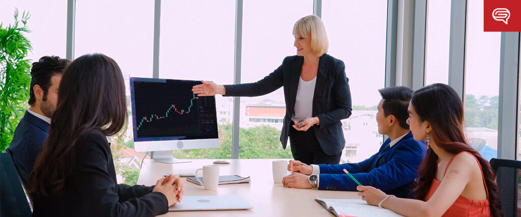 A group of five professionals, three women and two men, are gathered in a modern office. One woman is standing and pointing to a slide displaying a rising line graph. They are attentive, discussing the data. Large windows reveal an urban backdrop.