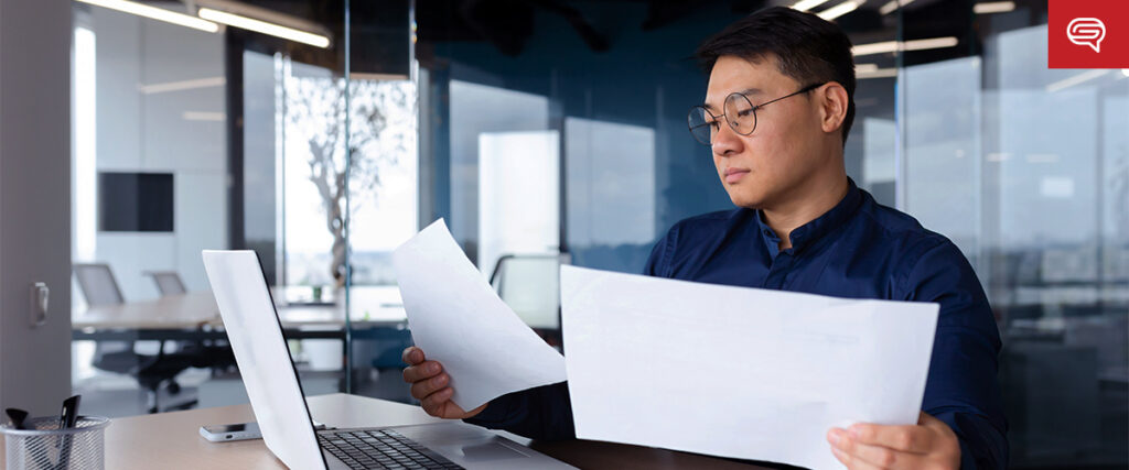 A man wearing glasses and a dark shirt sits at a desk in an office. He is looking at a piece of paper in his right hand while holding another sheet in his left hand. A laptop is open on the desk in front of him, displaying a PowerPoint presentation. The office has a large glass window.
