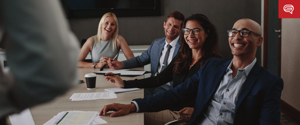 Four people in business attire sit at a conference table, smiling and laughing. Papers and a coffee cup are on the table. They appear to be participating in a meeting or presentation with PowerPoint slides. A person standing in the foreground is partially visible.